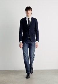 Tumed ülikond, mis on kohandatud sobivuseks, valge pidžaamapaita, must lips, tumedad teksad ja mustast nahast saapad, esitatud mudelil lihtsas keskkonnas.