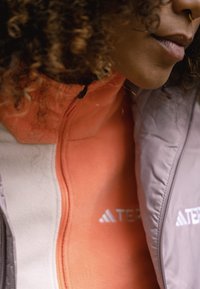Chaqueta de forro polar con una combinación de colores coral y gris, presenta cremalleras y un tejido texturizado. Logo de la marca visible en la parte frontal.