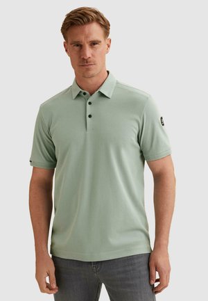 Mann mit kurzen hellbraunen Haaren, der ein hellgrünes kurzärmeliges Poloshirt und graue Jeans trägt, steht vor einfachem Hintergrund.
