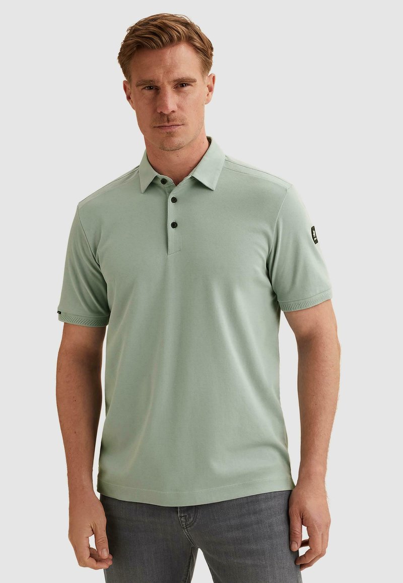 Homme aux cheveux courts châtain clair portant un polo vert clair à manches courtes et un jean gris, debout devant un fond uni.