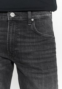 Persona vistiendo jeans de mezclilla negros con el bolsillo frontal visible y un botón de metal etiquetado "Lee Est. 1889" que cierra la cintura.