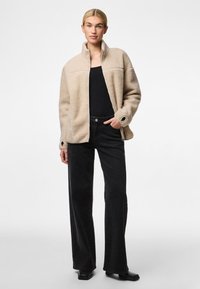 Jachetă din fleece bej cu guler înalt, având o textură interesantă și buzunare cu fermoar, asortată cu pantaloni negri evazați și botine.