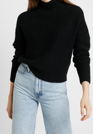 Žena oblečená v černém texturovaném pleteném svetru s dlouhými rukávy a světlých modrých džínech s vysokým pasem na jednobarevném pozadí.