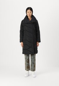 Svart boblejakke med bred krage, diagonal quiltet design og frontglidelås. Båret over mønstrede lyseblå bukser og hvite sneakers.