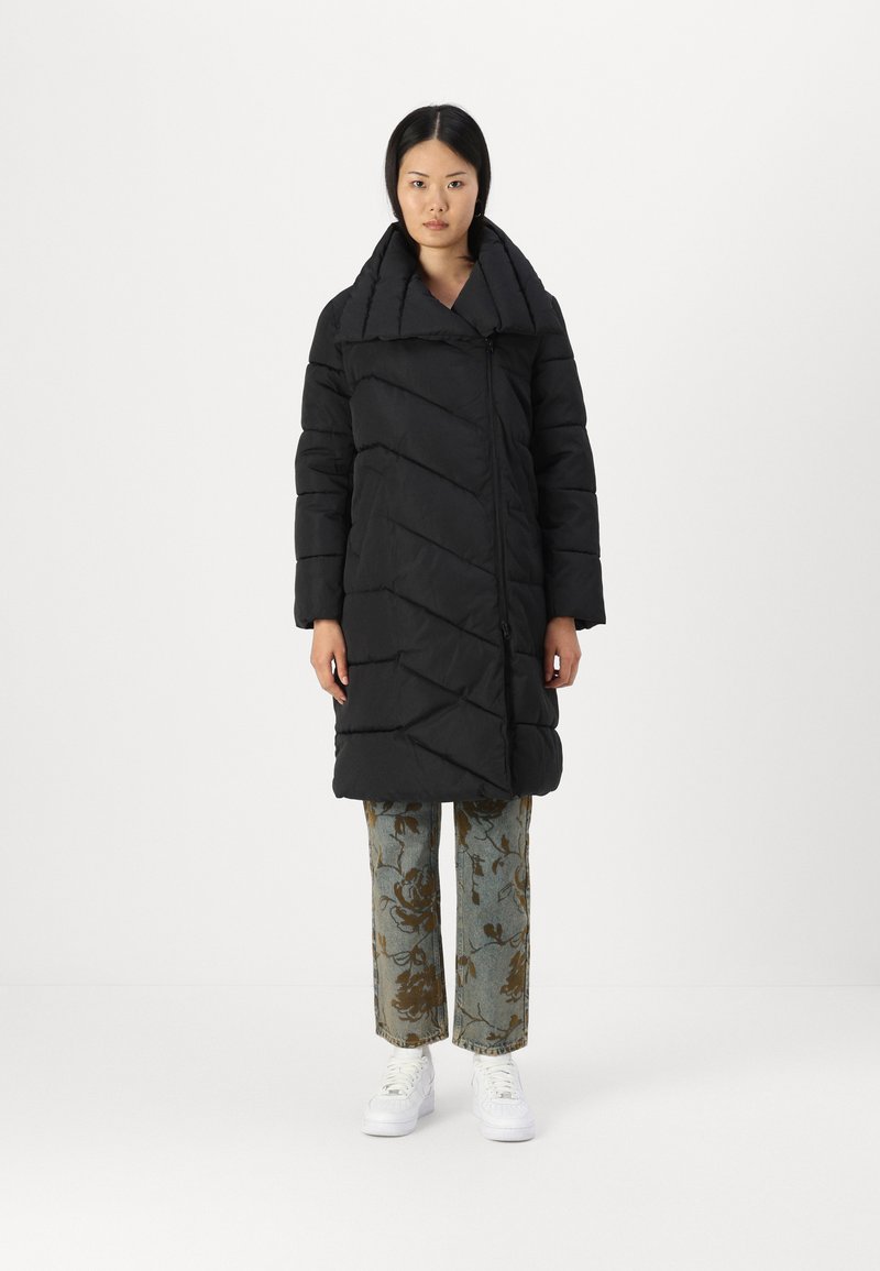 Svart boblejakke med bred krage, diagonal quiltet design og frontglidelås. Båret over mønstrede lyseblå bukser og hvite sneakers.