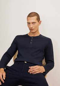 Camisa henley de manga larga en color navy con tapeta de tres botones y textura acanalada. Combinada con pantalones oscuros a juego. Modelo sentado en una silla.