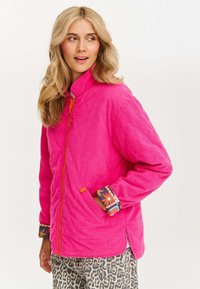 Różowa kurtka z kordurowej tkaniny, z wysokim kołnierzem, pomarańczowym zamkiem błyskawicznym i wzorzystymi, kwiatowymi mankietami. Posiada boczne kieszenie oraz luźny krój.