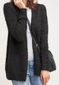Cardigan nero con texture pelosa, apertura frontale, maniche lunghe e una vestibilità rilassata. Indossato sopra una maglietta grigia e abbinato a jeans.