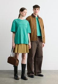 Δύο μοντέλα στέκονται δίπλα δίπλα φορώντας γαλάζια τοπάκια «Tommy Jeans»· η γυναίκα κρατά μια καφέ τσάντα, μια κίτρινη καρό φούστα και μαύρες μπότες.