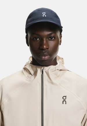 Jeune homme portant une veste zippée beige et une casquette bleu marine avec un logo blanc « on », faisant face à un fond gris uni.