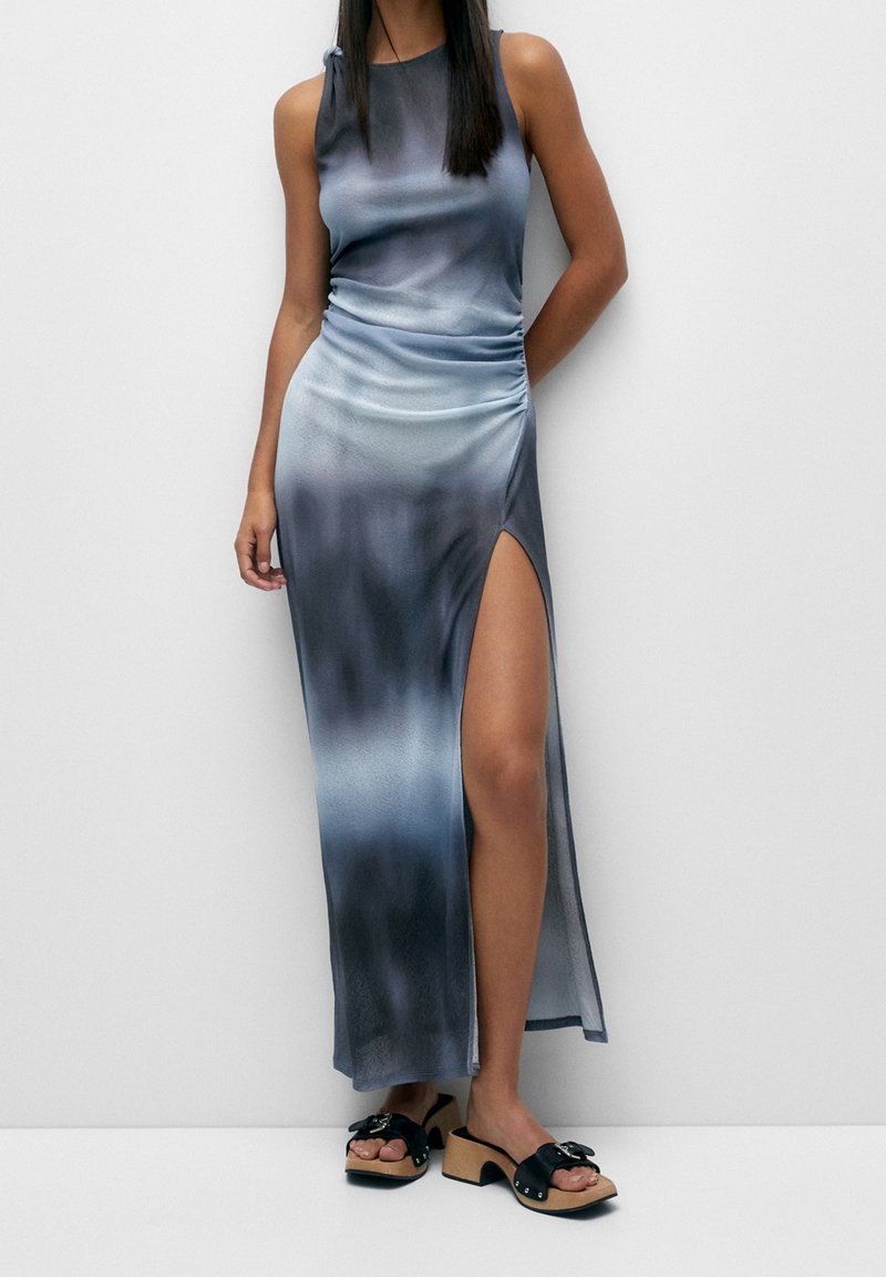 Femme portant une robe sans manches dégradée bleu-gris avec une fente haute à la cuisse et des sandales noires à talon bloc ouvertes à l’avant, debout contre un mur blanc.