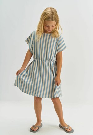 Fată tânără cu păr blond, purtând o rochie în dungi verticale albă și albastră și papuci gri, uitându-se în jos și ținând părțile rochiei.