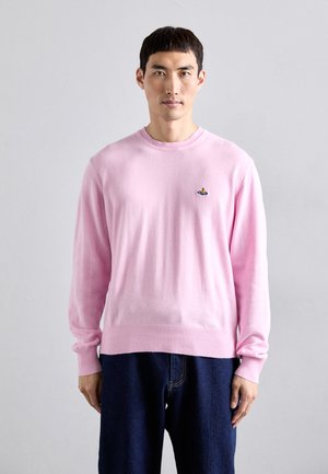 Mann in einem hellrosa Pullover mit kleinem besticktem Planetenlogo, kombiniert mit dunkelblauen Jeans, stehend vor einem schlichten hellen Hintergrund.