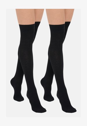 To par bein iført svarte ribbede knestrømper over kneet mot en ensfarget grå bakgrunn.