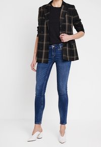 Blazer de cuadros negros con solapas, botones frontales; combinado con una camiseta negra y jeans skinny azules; unos mule blancos de tacón completan el conjunto.