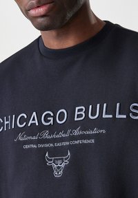 Černá mikina s kapucí s vyšitým nápisem "CHICAGO BULLS" šedou barvou, spolu s nápisem "National Basketball Association" a designem loga na přední straně.