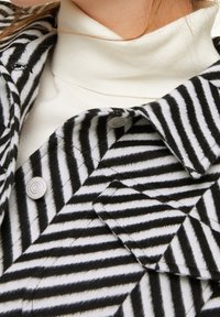 Sort-hvid stribet jakke med blød tekstur, udstyret med spids krave og knaplukninger, lagt over en hvid turtleneck.