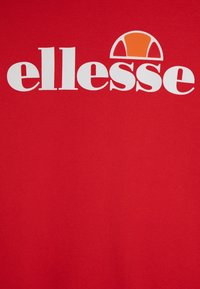 Camiseta de algodón roja con el logo blanco de "ellesse" y una gráfica redondeada en naranja y blanco encima del texto. Textura suave, diseño casual.