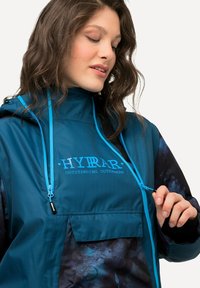 Una giacca teal e nera con cappuccio, cordini regolabili e tasca frontale, realizzata in tessuto testurizzato e con dettagli del logo.