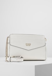 Borsa a tracolla Guess in pelle bianca testurizzata con tracolla in catena dorata e chiusura a flap, posizionata su un piedistallo bianco contro uno sfondo grigio.