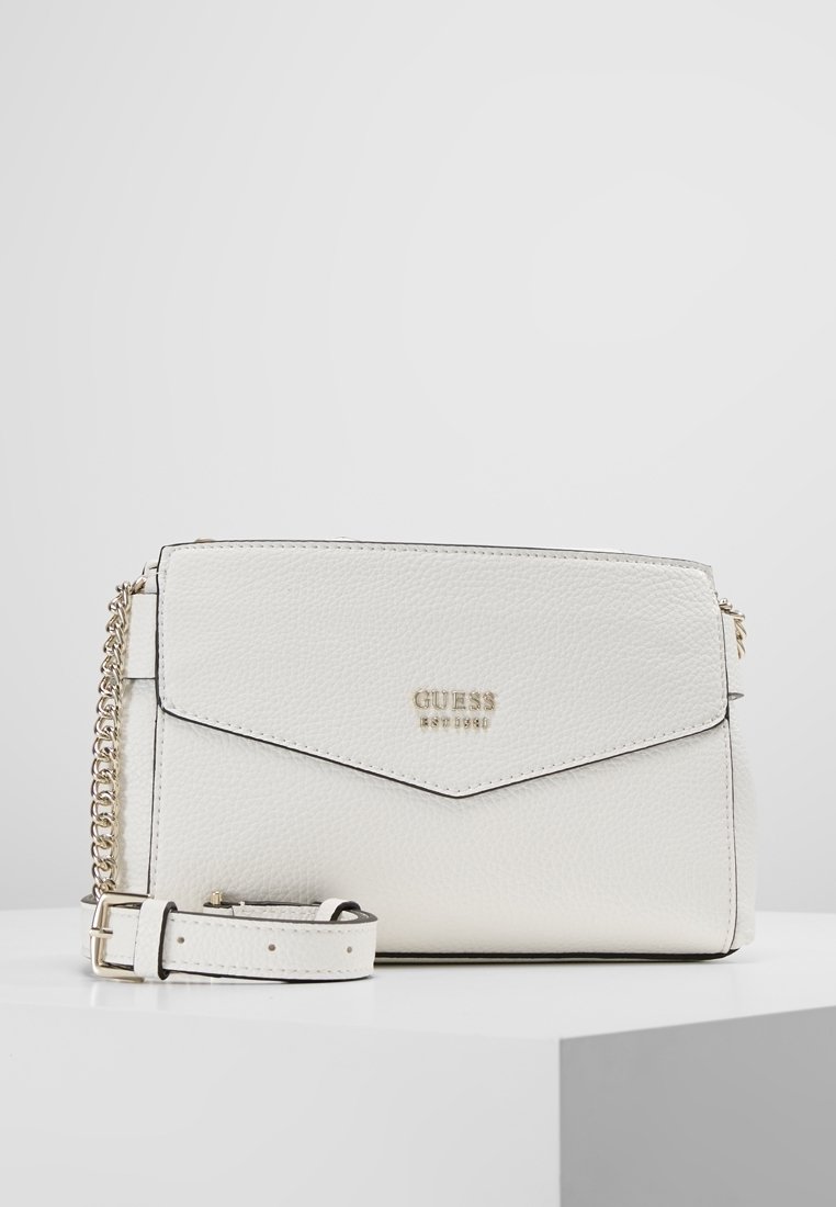 Borsa a tracolla Guess in pelle bianca testurizzata con tracolla in catena dorata e chiusura a flap, posizionata su un piedistallo bianco contro uno sfondo grigio.