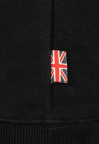 Black fabric with a textured finish featuring a small tag with a Union Jack design in red, white, and blue colors, sewn into the side seam.