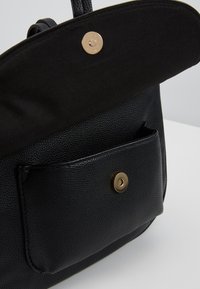 Svart texturerad handväska med lockstängning, som har en guldfärgad knapp och en ytterficka med en matchande guldfärgad knapp.