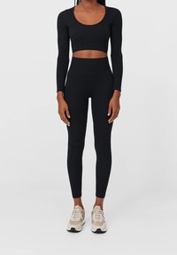 Zwarte cropped top met lange mouwen, gecombineerd met hoge zwarte leggings. Het materiaal lijkt zacht en aansluitend, aangevuld met beige sportschoenen.