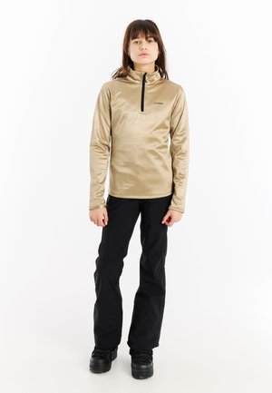 Hellbeige Zip-Up-Oberteil mit hohem Kragen, langen Ärmeln und kleinem Logo. Kombiniert mit schwarzen Hosen und klobigen schwarzen Schuhen.