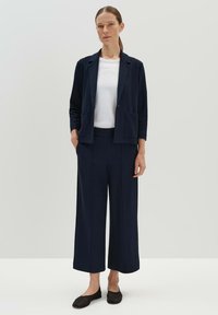 Marineblazer met twee voorkruiszakken, gecombineerd met een wit T-shirt en een wijde broek. Platte zwarte schoenen maken de outfit af. Zachte textuur.