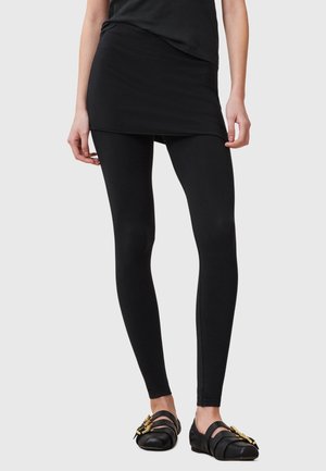 Leggings sportivi neri abbinati a una gonna sovrapposta nera coordinata, caratterizzati da un design aderente e una texture liscia. Accentuati da scarpe nere piatte.