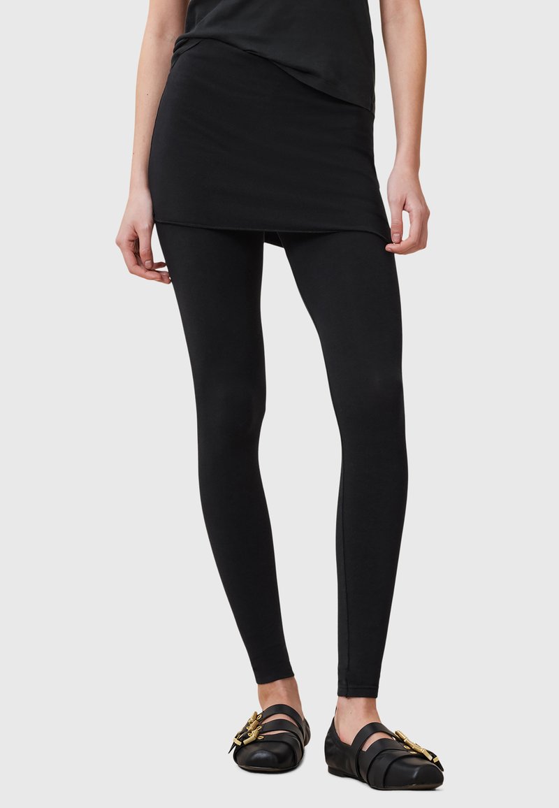 Leggings sportivi neri abbinati a una gonna sovrapposta nera coordinata, caratterizzati da un design aderente e una texture liscia. Accentuati da scarpe nere piatte.