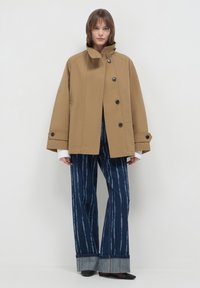 Frau in einer beige oversized Knopfjacket mit breiten Manschetten und blauen gestreiften weit geschnittenen Jeans, die vor einem schlichten weißen Hintergrund steht.