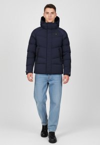 Granatowa kurtka puchowa z wysokim kołnierzem, suwakiem z przodu i dwoma bocznymi kieszeniami. W zestawie z jasnoniebieskimi dżinsami i czarnymi butami. Gładka tekstura.