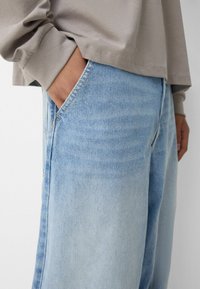 Hellblaue Jeans aus Denim mit einem lässigen Schnitt. Sie verfügen über eine sichtbare Tasche mit einem hellbraunen Stichdetail und einer glatten Stofftextur.