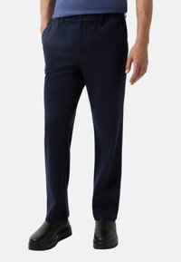 Marinblauwe broek met een losse pasvorm, elastische tailleband, voorplooien en zijzakken, gecombineerd met glanzende zwarte schoenen.