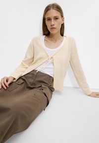 Różowa dziergana kardiganka z długim rękawem, otwartym przodem i zapięciem na związek; w połączeniu z białą koszulką na ramiączkach i brązowymi spodniami typu cargo. Gładka faktura.