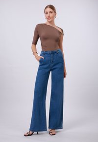 Bodysuit marrone aderente con design a spalla unica, abbinato a jeans in denim blu a gamba larga e vita alta. Modella che indossa tacchi neri con fasce.