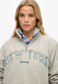 Sweat à zip gris avec "NEW YORK" en broderie verte et le texte "Superdry". Superposé sur une chemise à col bleu clair. Textile doux.