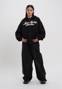 Sweatshirt preto com fecho éclair, apresentando texto em cursivo branco "Low Lights Studios", combinado com calças pretas largas e sapatos pretos brilhantes.