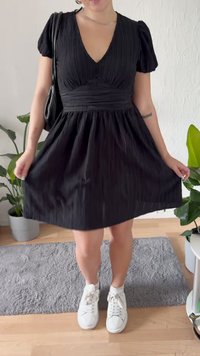 Μαύρο κοντό φόρεμα με V λαιμόκοψη, υφαντό ύφασμα και φουσκωτά μανίκια. Συνδυασμένο με λευκά αθλητικά παπούτσια, σε φόντο με εσωτερικά φυτά και γκρίζο χαλί.