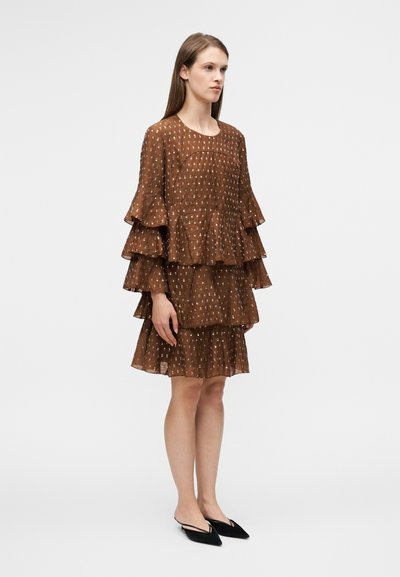 Robe marron à volants, composée de plusieurs couches, avec des manches volantées et un tissu texturé présentant de subtils motifs blancs. Associée à des ballerines noires pointues.