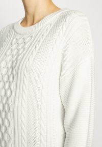 Biały dzianinowy sweter z teksturowanym wzorem warkocza na przodzie i prążkowanym, okrągłym dekoltem. Miękka tkanina o luźnym kroju i misternych wzorach.