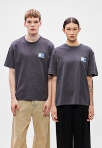 T-shirts cinzentas de algodão com gola redonda e mangas curtas, com um pequeno patch gráfico multicolorido no lado esquerdo do peito.