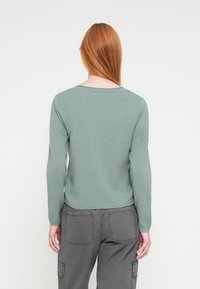 Μακρυμάνικη, ανοιχτοπράσινη μπλούζα από απαλό, λείο ύφασμα με στρογγυλή λαιμόκοψη και ριμπ μανσέτες, συνδυασμένη με γκρι παντελόνι cargo.