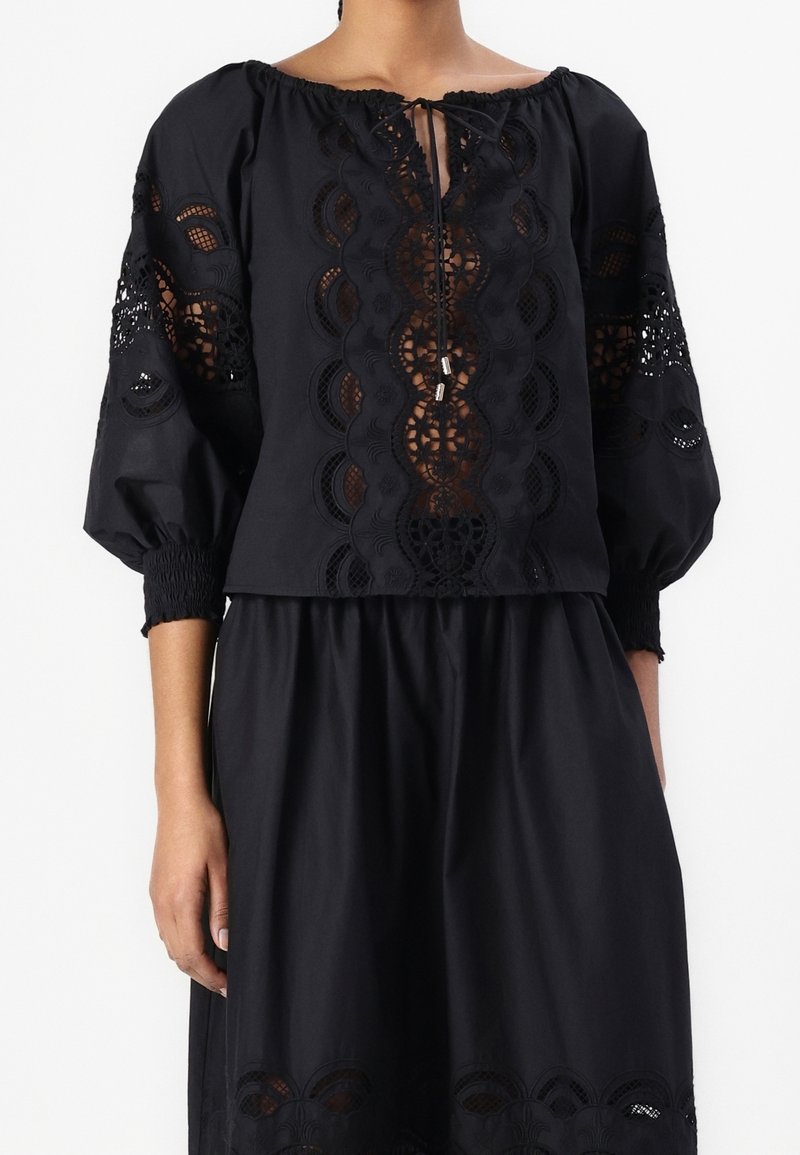 Blouse noire épaules dénudées avec des inserts en dentelle délicats sur le devant et les manches, associée à une jupe noire assortie avec un détail en dentelle à l’ourlet.