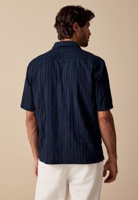 Marineblå kortærmet skjorte med lodrette strukturerede striber, knapper ned foran på kraven og en afslappet pasform, vist bagfra mod en neutral baggrund.