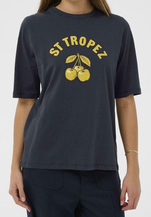 Vrouw draagt een donkerblauw T-shirt met gele tekst "ST TROPEZ" en een afbeelding van drie kersen met bladeren aan de voorkant.