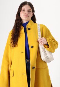 Cappotto di lana giallo brillante con colletto alla cinese, bottoni neri e tasche laterali. Il modello tiene una borsa bianca testurizzata con una tracolla decorata.