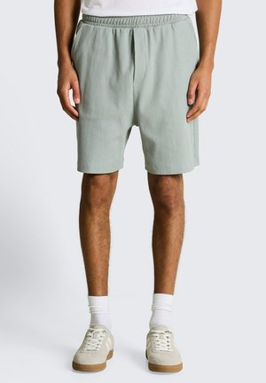 Mann trägt hellgrüne, gerippte Shorts mit elastischem Bund, weiße Socken, beige Turnschuhe und ein weißes T-Shirt, steht vor einem einfarbigen Hintergrund.